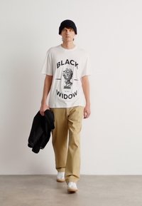 Hvitt T-skjorte med "BLACK WIDOW" grafikk, beige chinos, svart lue og hvite joggesko. Modellen holder en svart jakke. Minimalistisk bakgrunn.