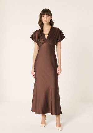 Femme portant une robe en satin marron à col en V avec manches courtes, un collier en chaîne dorée et des talons blancs à bout pointu, debout devant un fond uni.