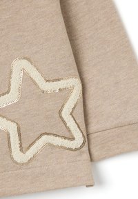Maglione beige con una texture morbida, caratterizzato da un design di stella contornato in tessuto bouclé dorato e crema con paillettes e cuciture dettagliate.