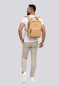 Zaino in pelle marrone chiaro con diverse tasche, forma rettangolare, spallacci imbottiti e maniglia superiore, indossato da una persona con pantaloni chiari.