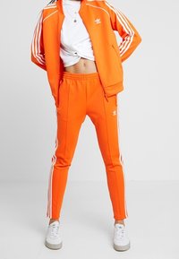 Orange idrottsoverall med vita ränder, bestående av en zip-up jacka och smalare byxor. Bärs med vita sneakers.