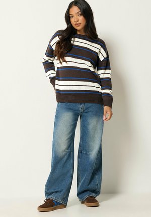 Femme debout vêtue d'un pull rayé marron, blanc et bleu, d'un jean bleu à jambes larges et de baskets marron sur un fond clair uni.