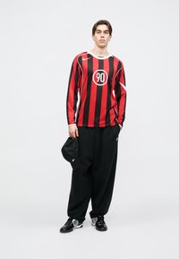 Μαύρη και κόκκινη ριγωτή μπλούζα μακρύ μανίκι με στρογγυλή λογότυπο και αριθμό "90," σε συνδυασμό με φαρδιά μαύρα παντελόνια και μαύρο καπέλο. Λιτές μαύρες παπούτσια.