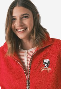 Czerwona polarowa kurtka z zamkiem błyskawicznym, z dodatkami w panterkę oraz haftem przedstawiającym Snoopy'ego. Nałożona na pasek z guzikami.