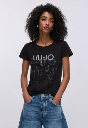 Giovane donna con capelli ricci che indossa una T-shirt nera con stampa grafica e jeans blu larghi, in piedi con le mani nelle tasche su uno sfondo semplice.