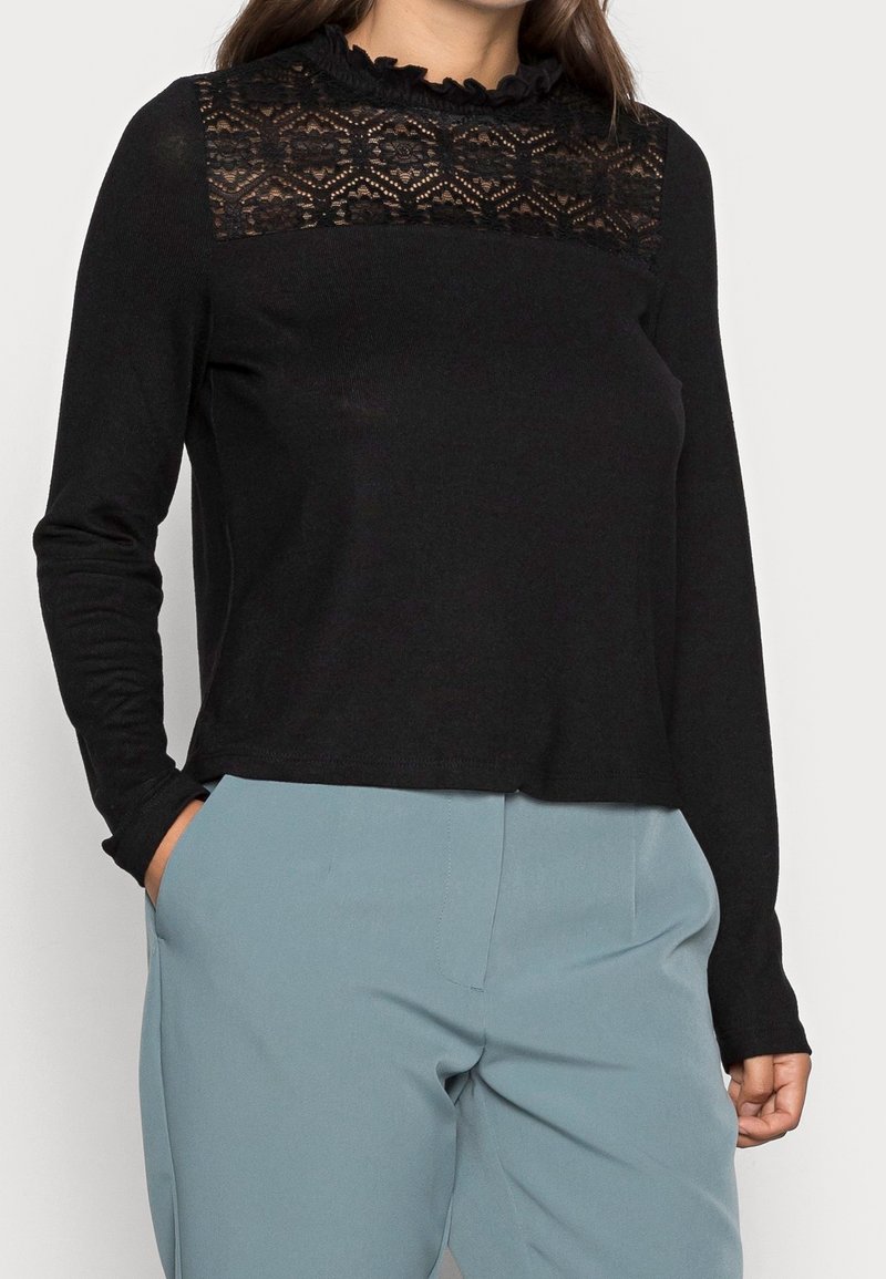 Femme portant un haut noir à manches longues avec des détails en dentelle au niveau du décolleté et un pantalon bleu clair, les mains dans les poches.