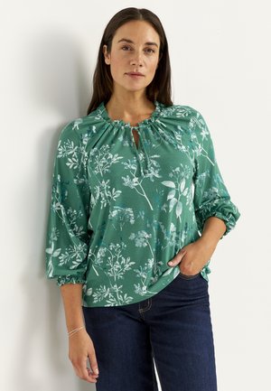 Frau mit langen dunklen Haaren, die eine grüne Bluse mit floralem Muster, gerafftem Ausschnitt und Dreiviertelärmeln trägt, kombiniert mit dunklen Jeans.