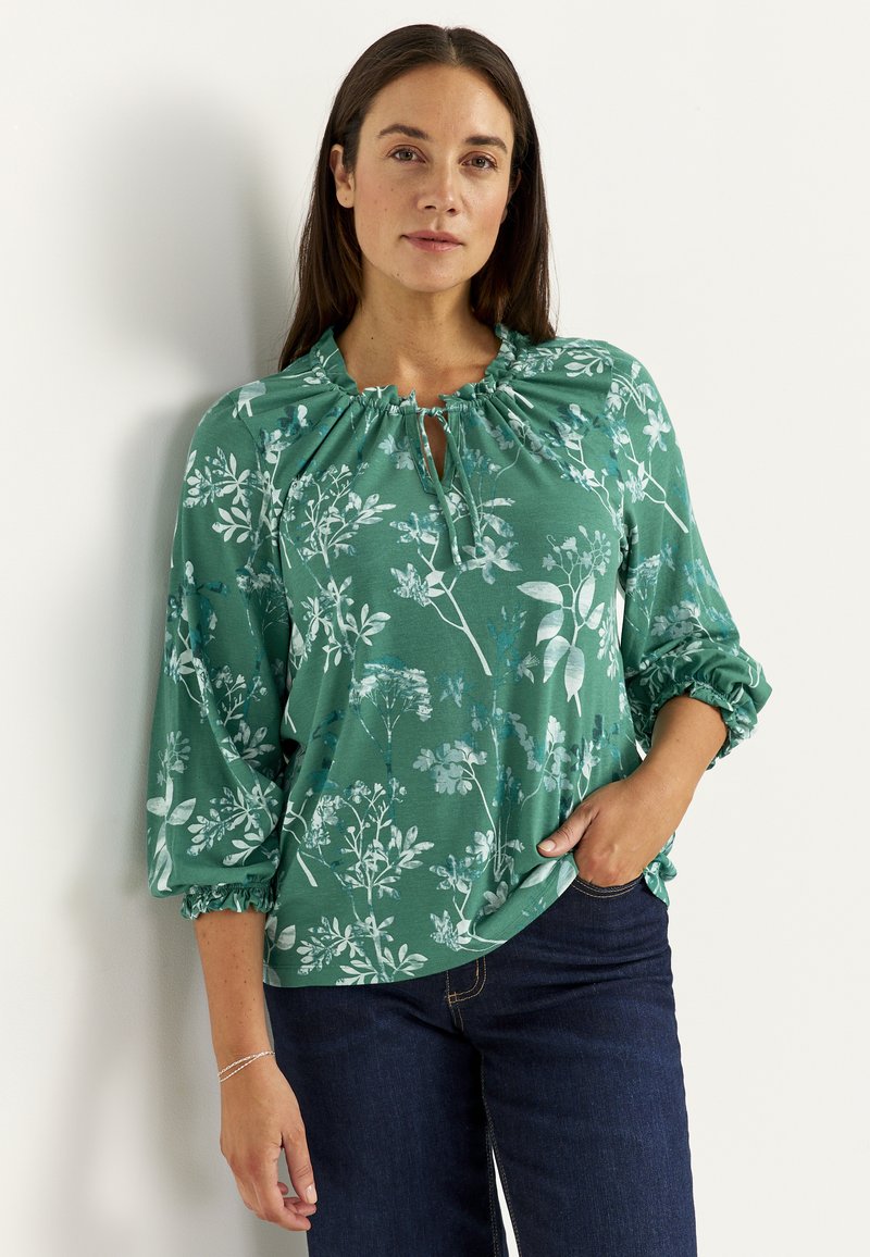 Kvinde med langt mørkt hår iført en grøn bluse med blomsterprint, samlet ved halsen og med trekvartlange ærmer, kombineret med mørke jeans.