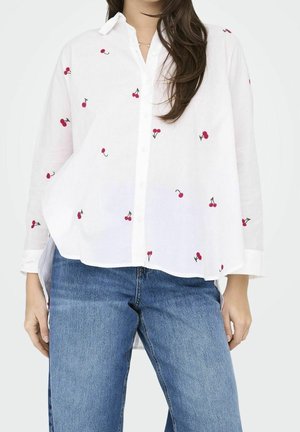 Femme portant une chemise blanche boutonnée avec de petites impressions rouges de cerises et un jean bleu, debout, les bras détendus le long du corps.