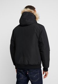 Sort isoleret jakke med hætte, der har en kant af kunstpels. Glat stof, elastiske ærmeafslutninger og linning med et enkelt design.