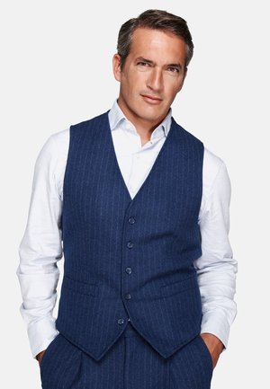 Man wearing a navy blue pinstripe vest and pants with a white dress shirt, standing with hands in pockets against plain background.