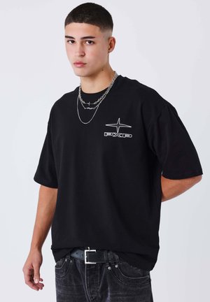 Camiseta oversized de algodón negro con mangas cortas, con un logo bordado en blanco "PXP" y un diseño de estrella. Llevada con cadenas de plata en capas.