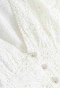 Witte stof met bloemige ajourborduursels, geplooide gedeelten en drie met stof beklede knopen in het midden.