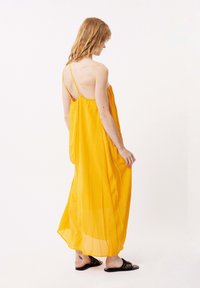 Langes, ärmelloses gelbes Kleid mit einem strukturierten Muster und dünnen Trägern, das bis zu den Knöcheln fällt, kombiniert mit schwarzen, offenen Sandalen.