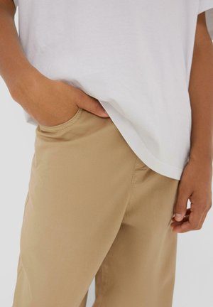 Persoon draagt beige broek met één hand in de zak en een wit overhemd deels ingestopt, tegen een effen achtergrond.