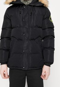 Mann trägt eine schwarze, gepolsterte Winterjacke mit einem mit Fell besetzten Kapuzen, durchgehendem Reißverschluss, Druckknöpfen, seitlichen Taschen und grünem gesticktem Aufnäher am Ärmel.