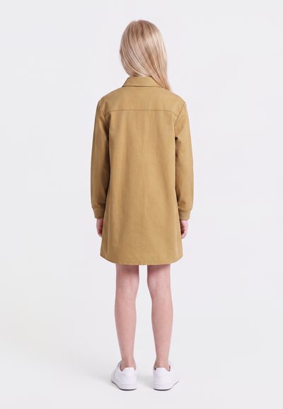 Enfant aux longs cheveux blonds portant une robe marron à manches longues et des baskets blanches, debout de dos sur un fond blanc uni.