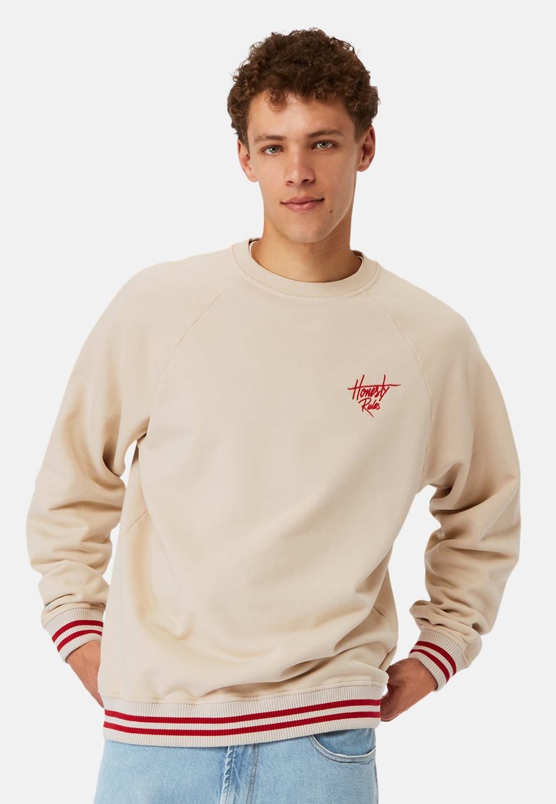 Beige sweatshirt med rød broderet logo, ribbede manchetter og kant med hvide og røde striber, blødt bomuldsmateriale, afslappet pasform.
