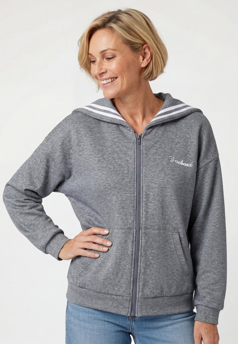 Femme souriante aux cheveux blonds courts portant un sweat à capuche gris zippé avec un col rayé et un jean bleu clair, main sur la hanche.