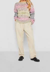 Pull tricoté multicolore avec des tons roses, jaunes et gris ; associé à un pantalon beige clair et des chaussures noires avec des accents dorés.