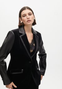 Blazer negro con cuerpo de terciopelo y mangas de cuero sintético, con solapa de satén, dos bolsillos frontales y cierre de un solo botón.