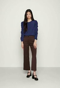 Mulher jovem com cabelo escuro e comprido, vestindo uma blusa quadriculada azul e roxa, calças largas castanhas e sapatos plataforma pretos, em pé tranquilamente.