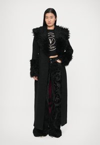 Dolgi črni plašč z umetnim krznom na ovratniku in manšetah, kombiniran s črnim kratkim topom in temnimi hlačami z vzorcem. Eleganten dizajn, teksturirana tkanina.