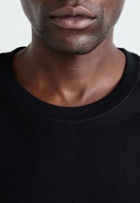 T-shirt en coton noir avec un col rond, texture lisse et des détails de couture visibles autour du col. Gros plan sur la zone du cou et de la bouche.