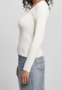 Biały prążkowany top z długim rękawem i dekoltem w serek, dopasowany, noszony z jasnoniebieskimi dżinsami z detalem kieszeni z tyłu.