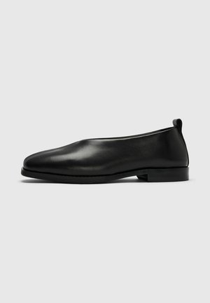 Zapato negro de piel sin cordones con tacón bajo y plano y punta redondeada, mostrado de perfil sobre fondo gris claro.