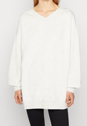 Vrouw die een loszittende witte lange mouw V-hals trui met geribbelde manchetten en een zwarte broek draagt, staande tegen een effen lichte achtergrond.