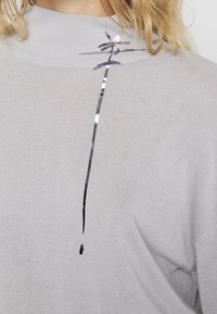 Ljusgrå stickad tröja med hög hals, prydd med ett reflekterande silveraccent diagonalt över bröstet. Mjuk textur och avslappnad design.