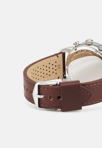 Brauner Lederarmband mit Edelstahlverschluss, versehen mit Perforationen und Kontrastnähten. Deutlicher Blick auf den transparenten Gehäuserücken.
