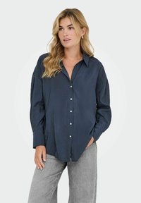 Donna bionda che indossa una camicia button-up larga di colore blu scuro e jeans grigi chiari, con una mano in tasca, mentre guarda leggermente di lato.