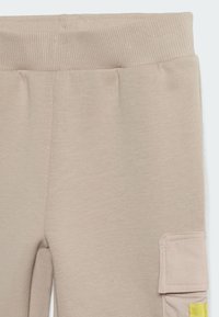 Beige jogger pants with a textured material, featuring a ribbed waistband and a side pocket with a yellow accent.