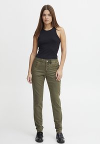 Zwarte mouwloze top gecombineerd met olijfgroene broek met vijf knopen en decoratieve zakken. De broek heeft een gladde textuur en een slanke pasvorm.
