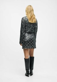 Robe noir pailleté à manches longues et à la coupe ajustée. Portée avec des bottes en cuir noir montantes, mettant en valeur une texture lisse et réfléchissante.