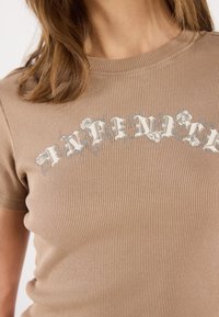 Šviesiai ruda, matinė T-marškiniai su trumpomis rankovėmis, puošti baltais gėlių motyvais, sukuriantys tekstūrinį raštą krūtinės srityje.