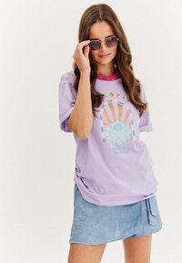 Lila oversized tričko s grafickým designem, který zobrazuje lotos a nebeské prvky. V kombinaci s světle modrou strukturovanou zavinovací sukní.