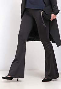 Svarta utsvängda byxor i slätt tyg, med åtalsande midja och en lätt glans. Kombinerad med svarta spetsiga klackar och en svart väska.