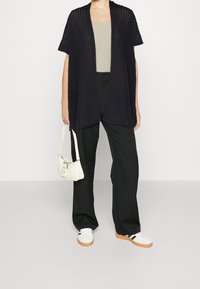 Öppenstickad svart cardigan med korta ärmar, lager på ett beige linne, kombinerat med vida svarta byxor och vita sneakers, hållande en liten vit väska.