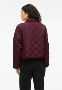 Burgundy quilted jacket with a cropped design, featuring a ribbed collar and smooth satin texture. Worn over black pants.