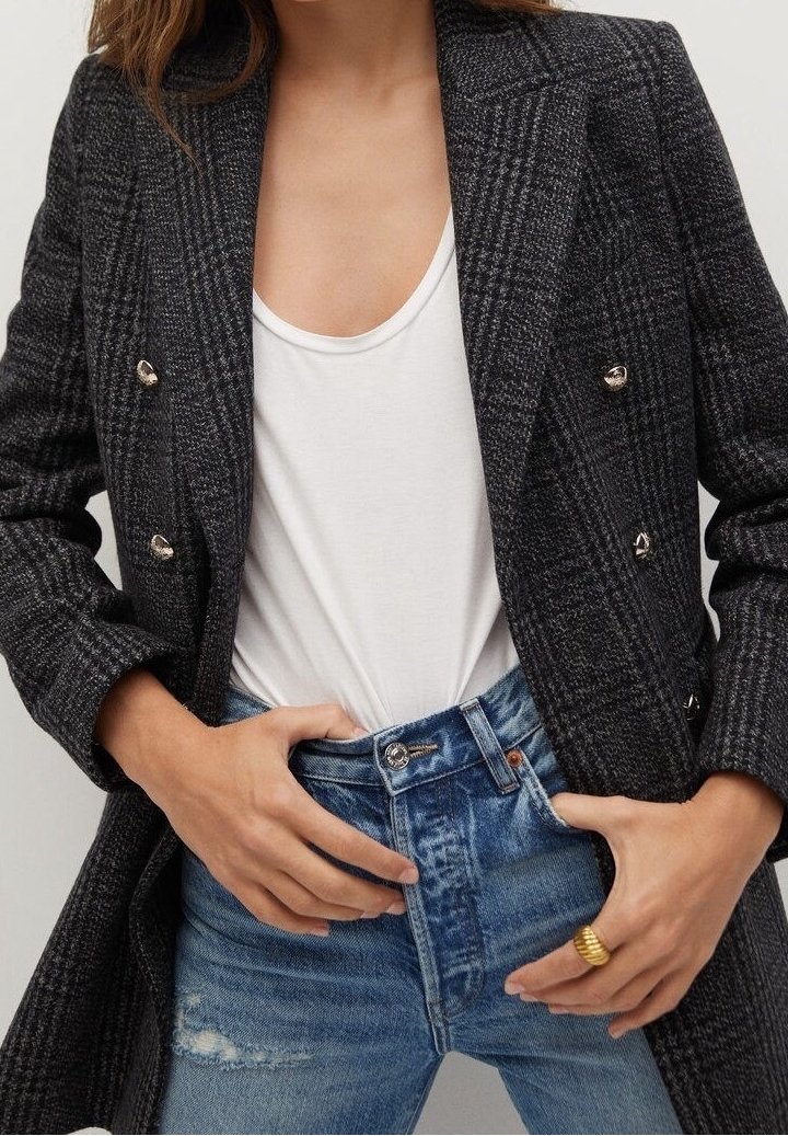 Femme portant un blazer croisé à carreaux gris foncé, un haut blanc à encolure ronde, un jean bleu taille haute et une bague en or à la main droite.