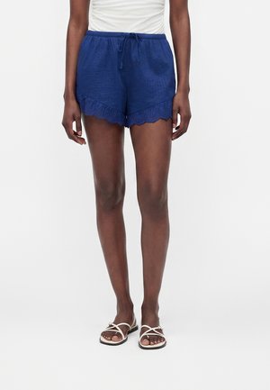 Frau trägt marineblaue Shorts mit Kordelzug und Spitzenbesatz und weiße Sandalen, steht vor weißem Hintergrund.