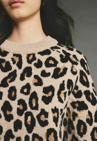 Femme portant un pull beige à imprimé léopard noir et marron, visible du menton au haut de la poitrine, sur un fond gris.