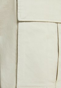 Beige tyg med en slät yta, som har en vertikal söm och en stor, platt ficka för användning.