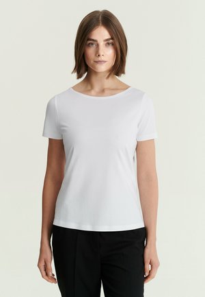 Jonge vrouw met kort bruin haar, gekleed in een effen wit T-shirt met korte mouwen en een zwarte broek, staand tegen een lichte achtergrond.