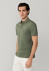 Giovane uomo con polo a maniche corte verde oliva con piccolo logo e pantaloni bianchi, in piedi davanti a uno sfondo grigio chiaro uniforme.