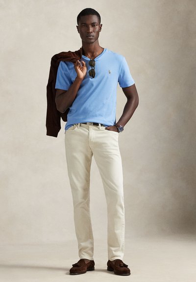 Šviesiai mėlyna trumpomis rankovėmis marškiniai, kreminės spalvos kelnės, rudi loaferiai ir ant peties užmestas rudas megztinis. Rankose akiniai nuo saulės.