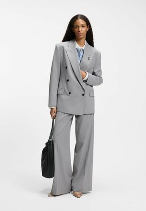 Femme aux longs cheveux bouclés portant un costume gris clair à double boutonnage, tenant un sac à main noir, debout devant un fond blanc uni.